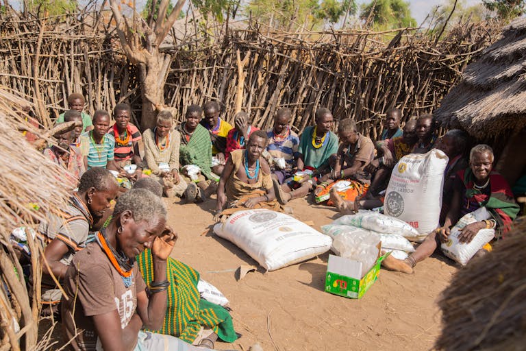 A group of African villagers seated outdoors surrounded by food supplies.